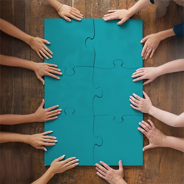 Le mani di molte persone attorno a un grande puzzle color teal, con le tessere già unite, su un tavolo di legno