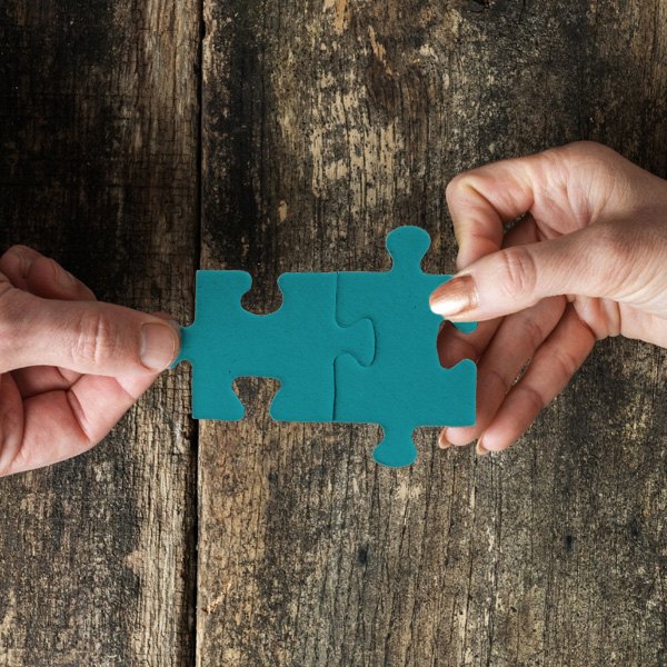 Due mani che avvicinano due tessere di puzzle color teal fino a farle combaciare, su un tavolo di legno